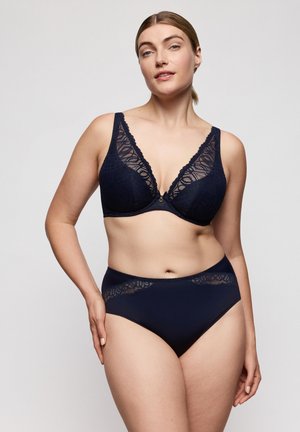 Vrouw staande met hand op heup, gekleed in een marineblauwe kanten beha en bijpassende hooggetailleerde onderbroek tegen een lichte achtergrond.