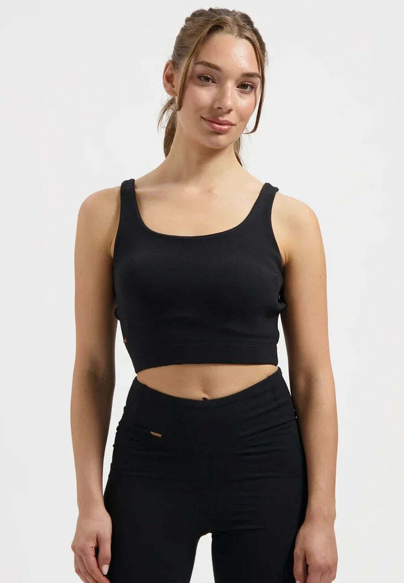 Junge Frau mit hellbraunen zurückgebundenen Haaren, trägt ein schwarzes, bauchfreies Tanktop und hoch taillierte schwarze Leggings, steht vor weißem Hintergrund.