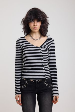 Νεαρή γυναίκα με σγουρά σκούρα μαλλιά, φοράει μακρυμάνικη μπλούζα σε μαύρο και γκρι ριγέ με ασύμμετρα κουμπιά και μαύρο παντελόνι με ασημένιες οπές.