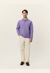 Pulover polo tricotat, texturat, de culoare purpurie, cu trei butoane, purtat cu pantaloni crem și pantofi negri. Modelul stă în fața unui fundal simplu.