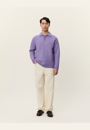 Fialový pletený polo svetr s texturou a třemi knoflíky, nošený s krémovými kalhotami a tmavými botami. Model stojí na jednobarevném pozadí.