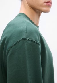 Sweatshirt verde em algodão com decote redondo, punhos canelados e uma textura suave. Apresenta detalhes de costura ao longo do ombro.