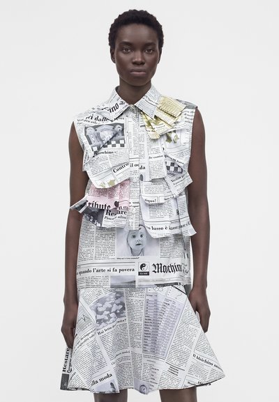 Γυναίκα που φοράει αμάνικο φόρεμα με γιακά, με τύπωμα εφημερίδας και λεπτομέρειες με στρωμένες βολάν σε απλό φόντο.