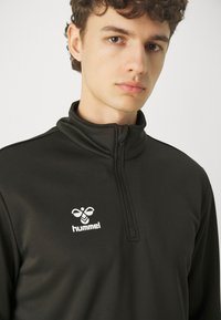 Schwarzer Reißverschluss-Hoodie mit hohem Kragen, mit einem weißen "hummel"-Logo auf der Brust. Glatte Textur, taillierte Passform, minimalistischer Stil.