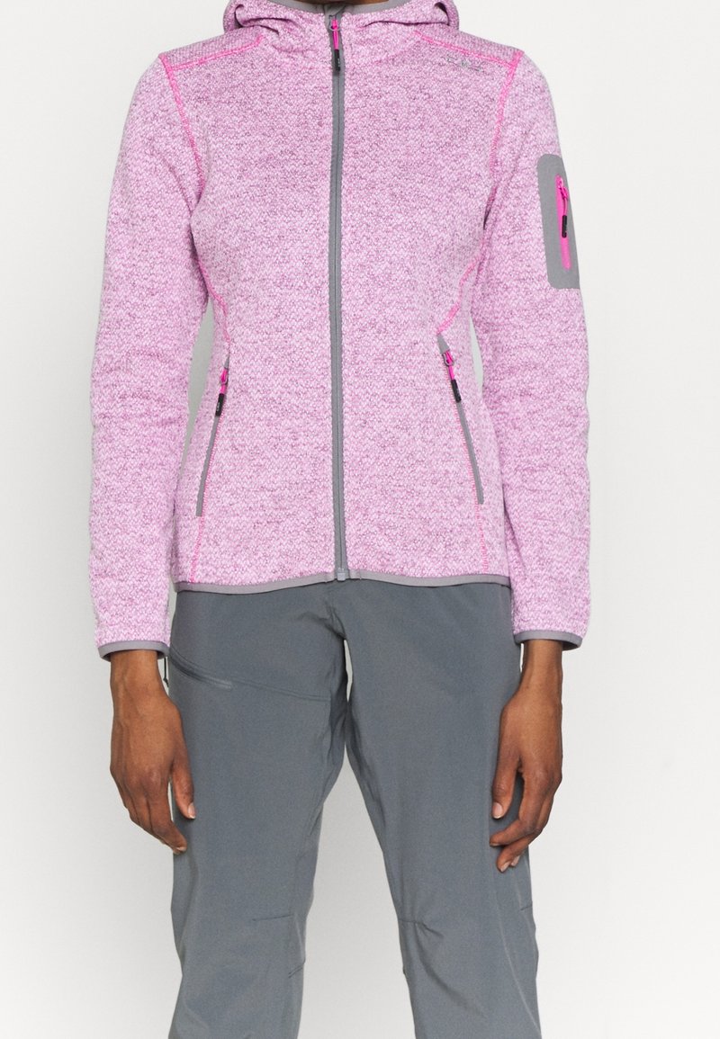 Personne portant une veste à capuche rose zippée avec des poches zippées sur les côtés et les manches, associée à un pantalon gris.