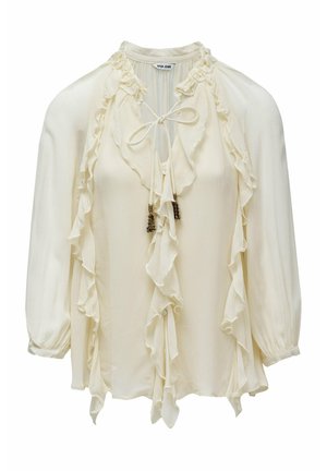 Blouse crème à manches longues, bordure volantée le long du devant et de l'encolure, et lien avec perles décoratives au niveau du cou.