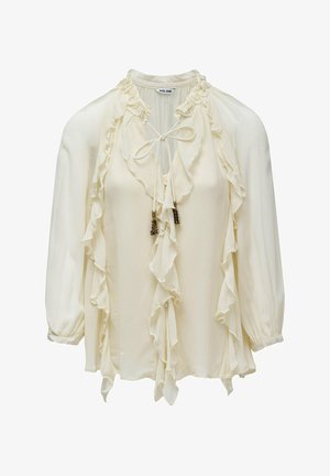 Blouse crème à manches longues, bordure volantée le long du devant et de l'encolure, et lien avec perles décoratives au niveau du cou.