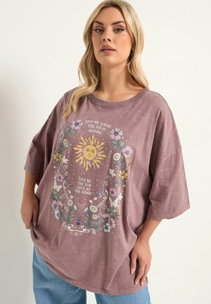 Frau trägt ein locker sitzendes mauvefarbenes T-Shirt mit Sonne, Blumen, Schmetterlingen und dem Text „Finde mich dort, wo die Sonne scheint“ und „Lebe nach der Sonne, liebe nach dem Mond.“