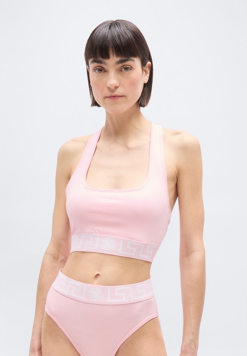 Γυναίκα που φοράει αθλητικό σουτιέν σε ανοικτό ροζ χρώμα και εσώρουχο ψηλόμεσο με ελαστικές ταινίες σε λευκό με γεωμετρικό μοτίβο, στέκεται μπροστά σε απλό γκρι φόντο.