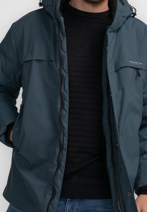 Mann trägt dunkeltürkisfarbenen Kapuzenjacke über schwarzem Pullover und blauen Jeans, mit Händen in den Jackentaschen, sichtbarer Reißverschluss und Taschen.