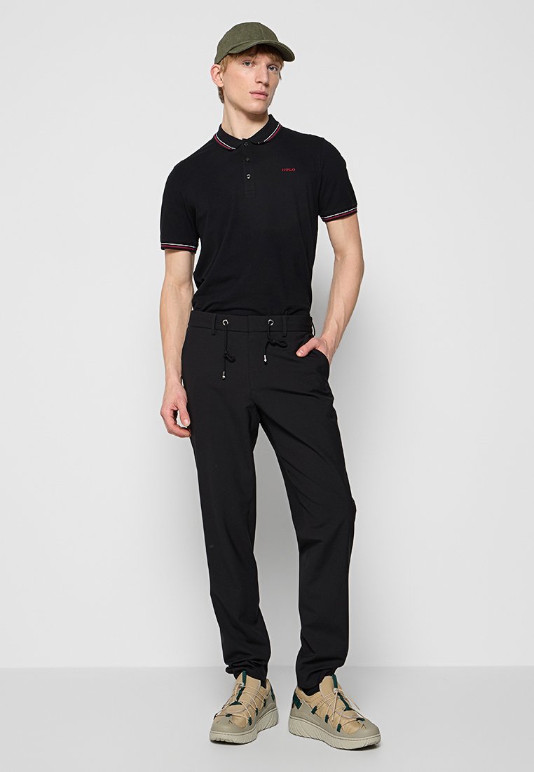 Polo shirt noir avec un col rayé rouge et blanc, associé à un pantalon noir avec une taille à cordon. Les chaussures sont beiges avec des accents verts.