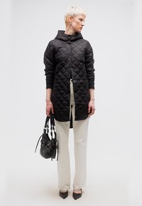 Γυναίκα με κοντά ξανθά μαλλιά που φοράει μαύρο καπιτονέ παλτό, λευκό παντελόνι, μαύρα μυτερά παπούτσια, κρατώντας μια μαύρη τσάντα, στέκεται σε λευκό φόντο.