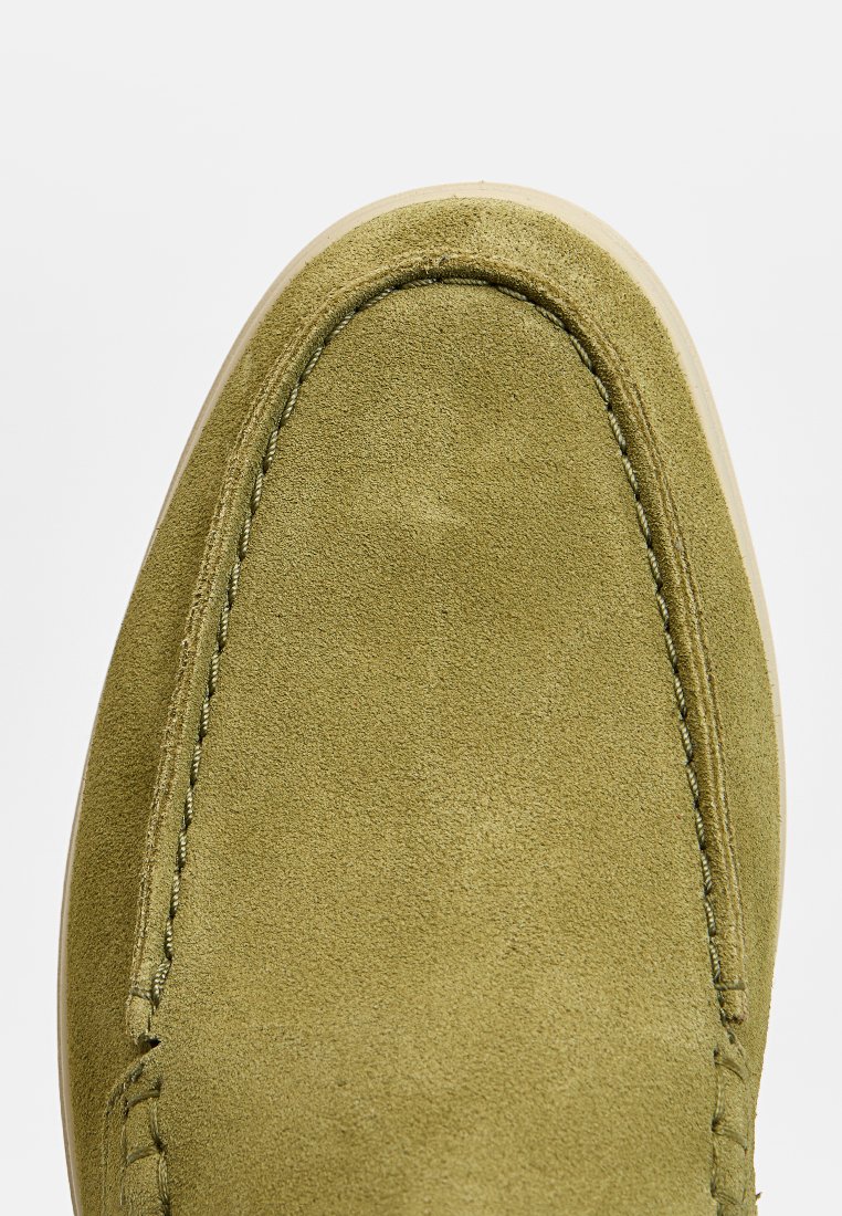 Chausson en suède vert avec un bout arrondi, semelle beige et détails de couture visibles le long du périmètre. Texture lisse dans l'ensemble.
