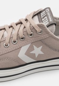 Beige kankaan lenkkitossu valkoisella tähden logolla, metalliset nauha-aukot ja beige nauhat. Varustettu valkoisella ja mustalla raidallisella kumipohjalla.