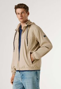 Béžna lahka jakna s kapuco, elastičnimi manšetami in pasom. Ima kontrastne vrvice za zategovanje in dva sprednja žepa. Oblečena čez modro srajco.