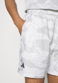 Vita shorts med ett ljust grått abstrakt mönster, elastisk midja, sidofickor och svart Adidas-logga på nederkanten.