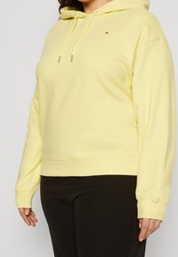 Osoba w jasnożółtej bluzie z kapturem i czarnych spodniach, pokazująca tors i ramiona na jednolitym jasnym tle.