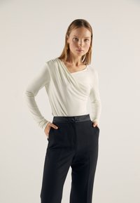 White long-sleeve top with a draped design and a square neckline, paired with high-waisted black trousers featuring side pockets.