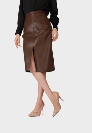 Donna che indossa una gonna in pelle marrone al ginocchio con uno spacco frontale, una maglia nera a maniche lunghe e scarpe beige con tacco alto, in piedi con le gambe incrociate.