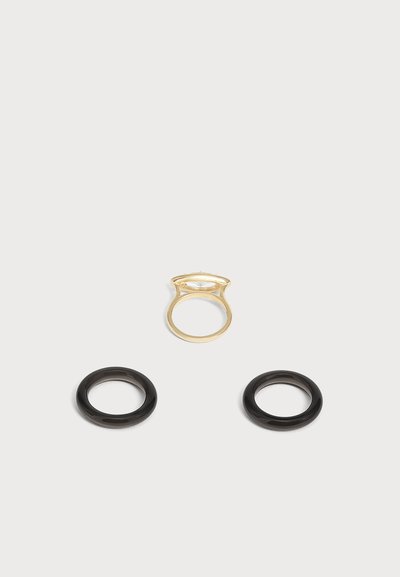 Une bague en or avec une pierre claire et deux bagues noires lisses et simples sur un fond gris clair.
