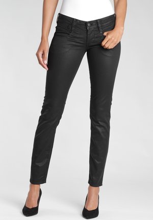 Nainen, jolla on mustat, kapeat nailonpinnoitetut farkut ja mustat korkokengät, seisoo yhdellä kädellä taskun lähellä neutraalia taustaa vasten.