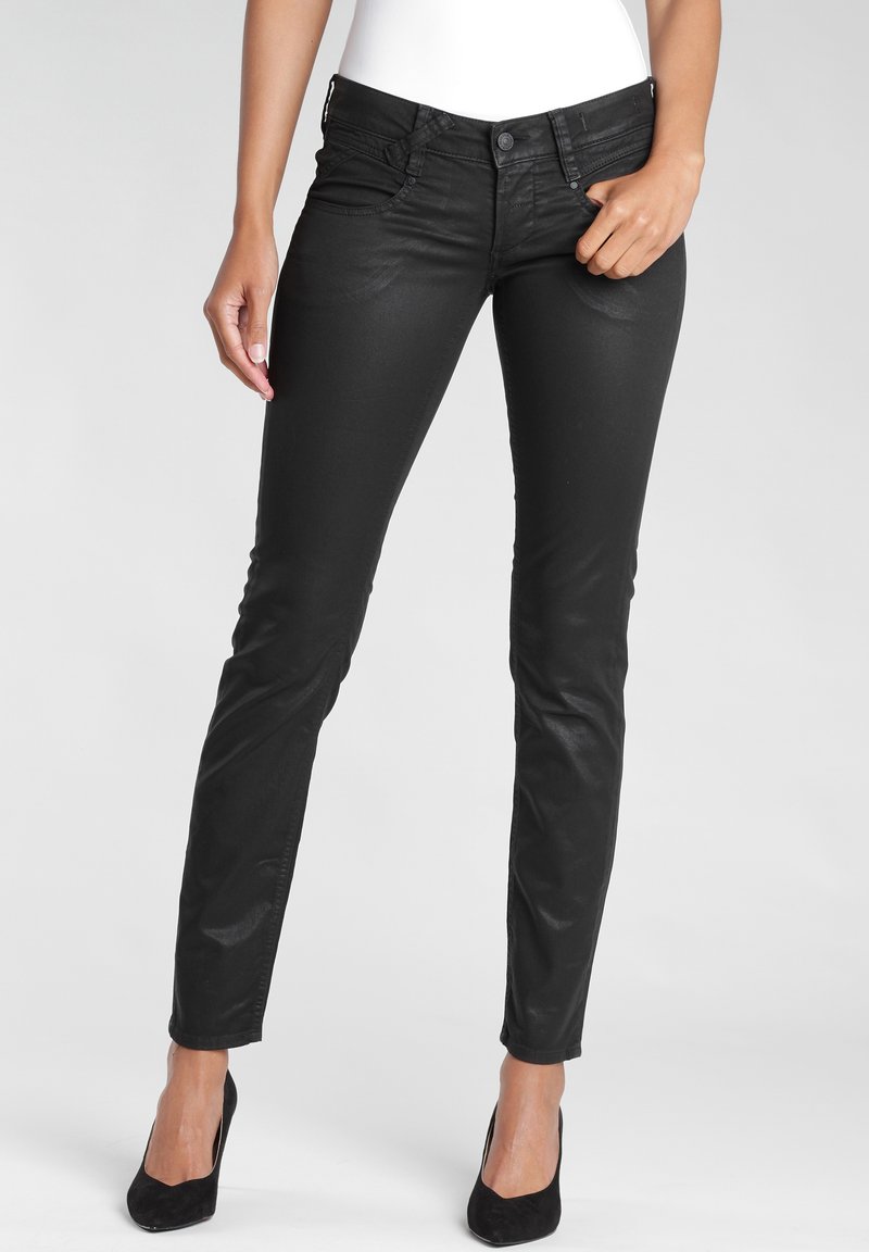 Nainen, jolla on mustat, kapeat nailonpinnoitetut farkut ja mustat korkokengät, seisoo yhdellä kädellä taskun lähellä neutraalia taustaa vasten.