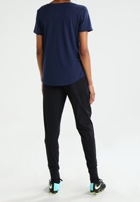 Person i en marinblå kortärmad skjorta, svarta byxor och svarta träskor med ljusblå klackar, som står vänd bort från kameran mot en vit bakgrund.