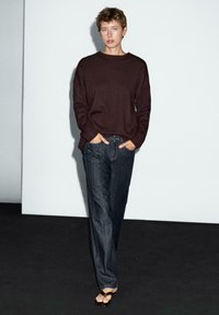 Bordeauxrotes Sweatshirt mit oversized Passform, kombiniert mit dunkelblauen weit geschnittenen Jeans. Das Model trägt dünne schwarze Sandalen und steht vor einem hellen Hintergrund.