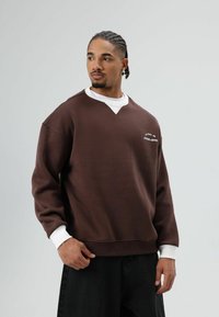 Bruine sweatshirt met een witte ronde hals en manchetten, met een V-vormige halslijn en tekstdetails op de borst. Zachte, gestructureerde stof.