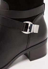 Gros plan d’une botte noire en cuir à la cheville avec un talon bloc bas et une boucle décorative argentée autour de la partie supérieure.
