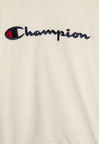 Kremowa bluza z kapturem z haftowanym logo "Champion" w odcieniach navy i czerwonym. Miękka faktura, luźny krój, minimalistyczny design.