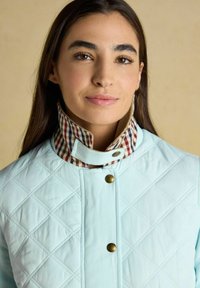 Mujer con cabello largo y oscuro que lleva una chaqueta acolchada azul claro con cuello a cuadros y botones de presión de latón, fondo neutro.