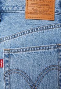 Blå denimtyg med en läderetikett med prägling. Märkbar röd Levi's-flik och kontrasterande sömmar längs fickorna.