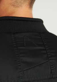 Achterkant van de nek van een persoon met kort haar, gekleed in een zwarte jas met een geribde kraag en een gestikt ontwerp op de bovenrug.