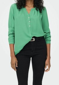 Blusa verde con bottoni, scollatura a V, maniche lunghe, tessuto texturizzato e orlo curvo, abbinata a pantaloni neri e una cintura slim.