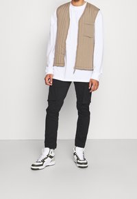 Beige gewatteerde vest met een voorkantzak over een witte langemouwenshirt, gecombineerd met zwarte cargobroeken en witte sneakers met groene accenten.