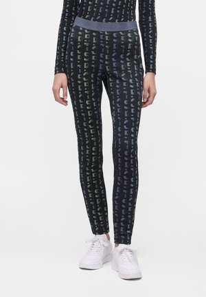 Femme portant des leggings noirs et un haut assorti avec des motifs de texte verticaux gris et colorés, associés à des baskets blanches à lacets.