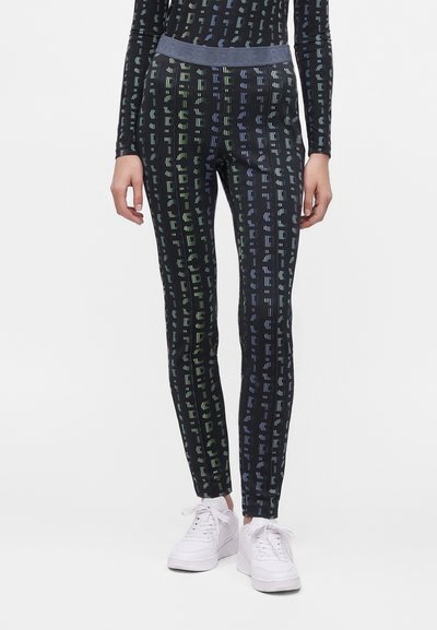 Femme portant des leggings noirs et un haut assorti avec des motifs de texte verticaux gris et colorés, associés à des baskets blanches à lacets.