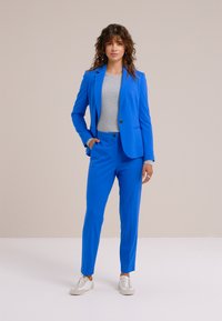 Svijetloplava uska sako jakna s odgovarajućim hlačama, uparena sa svijetlosivom majicom i bijelim tenisicama. Glatka tkanina, uski kroj i minimalni detalji.