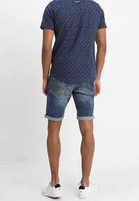 Męska granatowa koszulka z małym wzorem palm, zestawiona z deniowymi szortami z podwiniętym mankietem w lekko wyblakłym niebieskim odcieniu oraz białymi sneakersami.