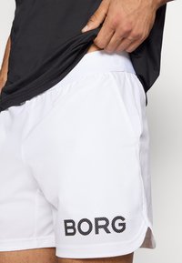 Calções atléticos brancos feitos de um tecido leve, apresentando um logótipo preto "BORG". Os calções têm uma textura suave e bolsos laterais.