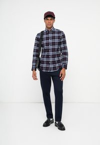 Camicia a quadri in blu navy, rosso e bianco; maniche lunghe con polsini a bottoni. Abbinata a pantaloni blu navy e scarpe nere, mentre porta una borsa a tracolla nera.