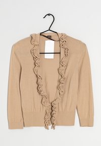 Cardigan beige avec un devant ouvert à volants, des manches longues et des poignets et un ourlet côtelés. Le tissu semble doux avec une texture lisse. Étiquette attachée.