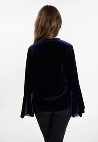 Top in velluto blu navy con maniche svasate, texture liscia e un semplice scollo a giro. Indossato con capi inferiori neri.