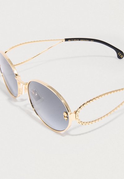 Occhiali da sole in metallo dorato con lenti grigie ovali, aste decorative testurizzate e un accento con logo vicino alla lente. Design elegante e unico.