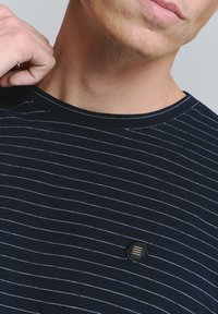 Marineblauwe T-shirt met witte horizontale strepen, ronde hals en een klein geborduurd hexagonaal logo op de borst. Zachte stoftextuur.