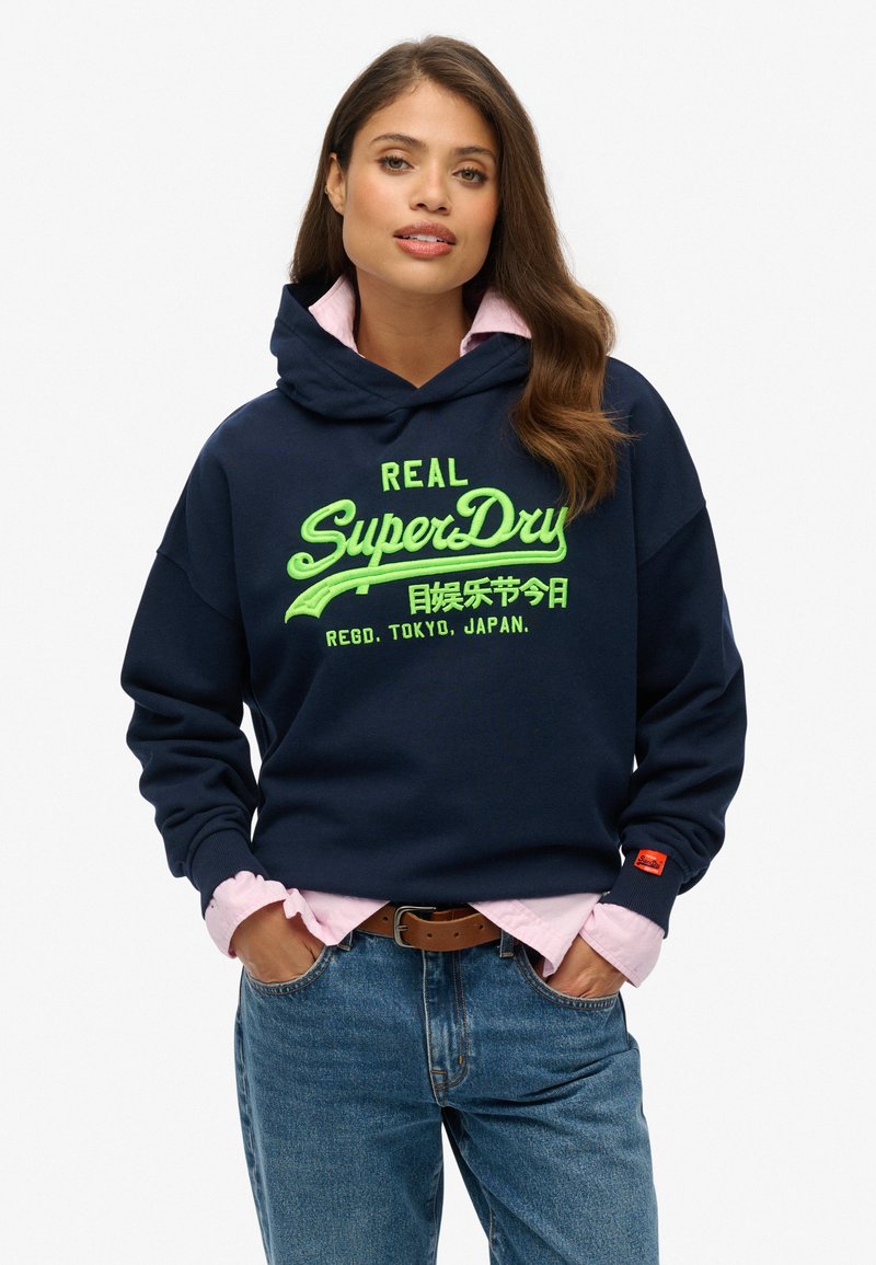 Sudadera con capucha de color azul marino con texto de logo en verde brillante, superpuesta a una camisa de cuello de color rosa claro, combinada con jeans azules.