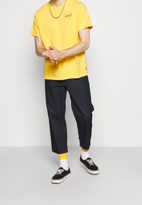 T-shirt di cotone gialla con logo Levi's, abbinata a pantaloni in denim scuro. Sneakers nere con suola bianca e calzini con accentuazione gialla.