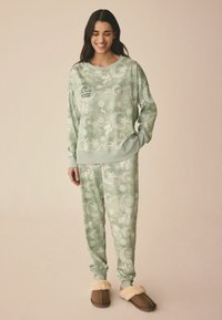 Grøn mønstret loungewear sæt med måner og stjerner; blødt stof, afslappet pasform og ribbede manchetter. Sammen med brune slippers.