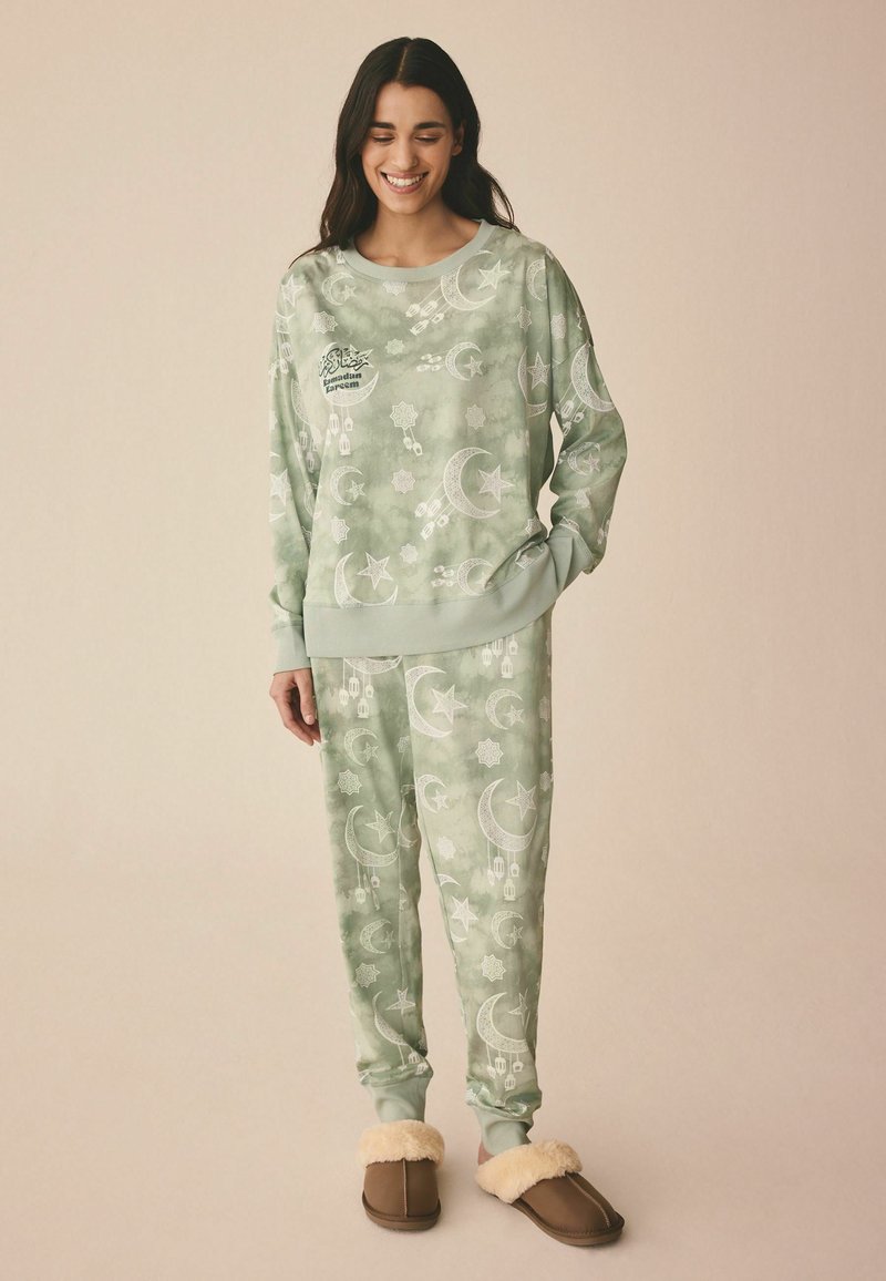 Grøn mønstret loungewear sæt med måner og stjerner; blødt stof, afslappet pasform og ribbede manchetter. Sammen med brune slippers.